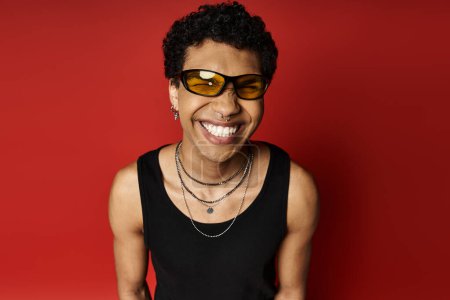 Foto de Hombre afroamericano guapo con gafas de sol elegantes sonríe contra un llamativo telón de fondo rojo. - Imagen libre de derechos