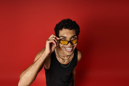 Téléchargez les photos : Un bel homme afro-américain porte avec confiance des lunettes de soleil sur un fond rouge audacieux. - en image libre de droit
