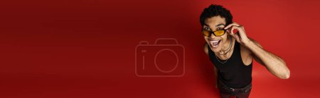 Handsome African American man with sunglasses stands in front of bold red backdrop.