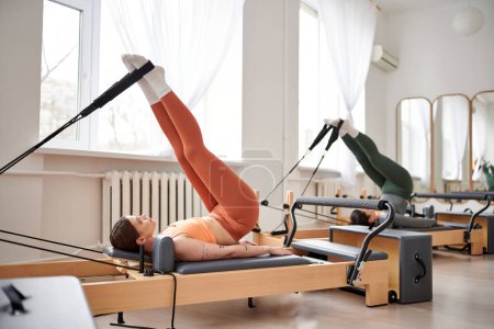 A sporty woman in an orange top stretches her back during a Pilates lesson.
