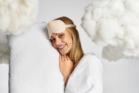 A dreamy blonde woman in cozy pyjamas holds a pillow, surrounded by fluffy clouds.