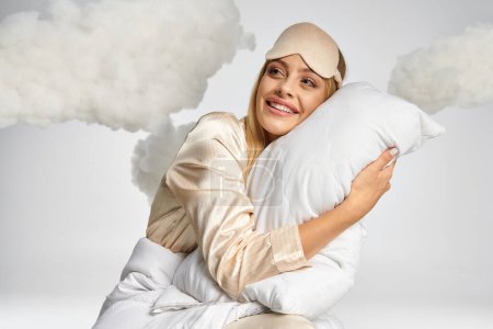 Verträumte blonde Frau im kuscheligen Pyjama sitzt zwischen Wolken mit einem Kissen.