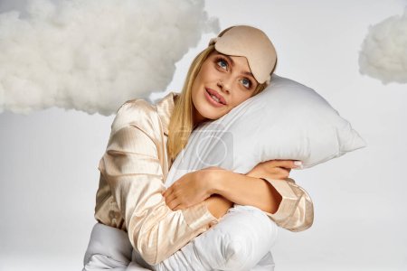 Blonde Frau im kuscheligen Pyjama mit flauschigem Kopfkissen umgeben von Wolken.