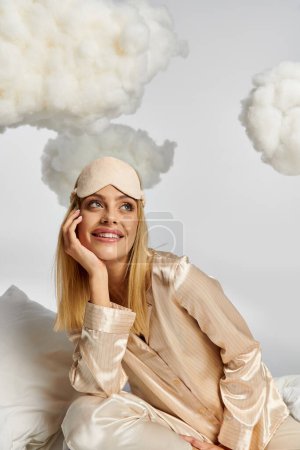 Eine verträumte blonde Frau im kuscheligen Pyjama sitzt auf einem weißen Kissen zwischen flauschigen Wolken.