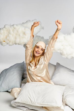 Eine verträumte blonde Frau im kuscheligen Pyjama sitzt zwischen Kissen auf einem Bett.
