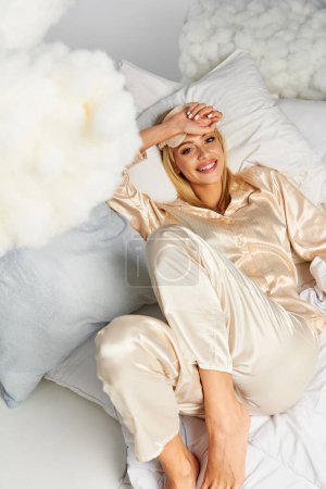 Foto de Una mujer rubia de ensueño en pijamas acogedores pone encima de una cama al lado de almohadas. - Imagen libre de derechos