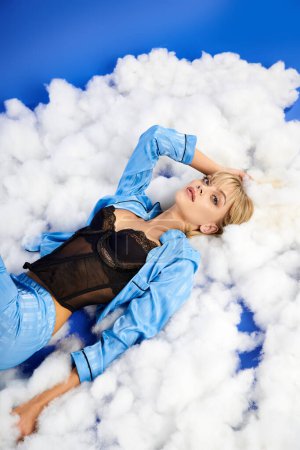 Une femme blonde captivante en tenue vibrante repose sur une pile de nuages pelucheux contre un ciel bleu.