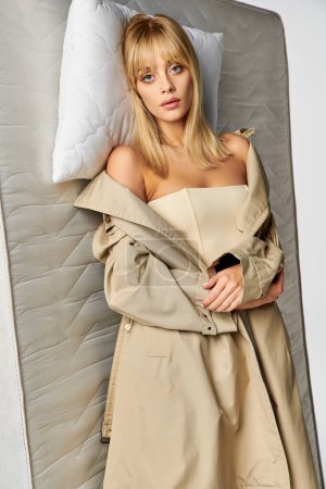 A stylish woman in a trench coat lounging against a pillow and matress.