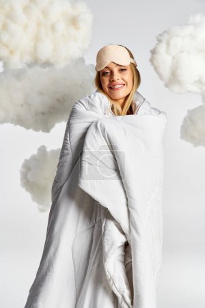 Photo for Blonde woman in cozy pyjamas, draped in white blanket, posing in fluffy clouds. - Royalty Free Image