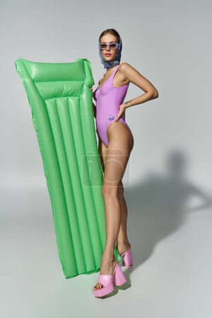 Fashionable woman in purple swimsuit holding inflatable mattress.