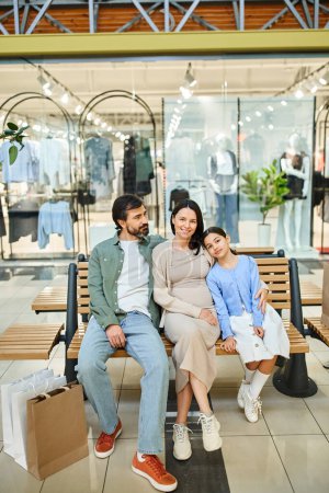 A happy family, enjoying a shopping weekend, sits together on a bench in a bustling mall, sharing a moment of togetherness.