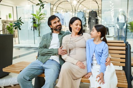 Eine schwangere Frau und ihre Tochter genießen einen Moment zusammen mit einem Mann auf einer Bank in einem Einkaufszentrum während eines Wochenendausflugs.