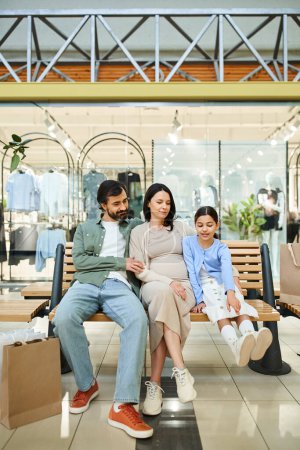A happy family, dressed casually, sits together on a bench in a bustling shopping mall, taking a break from their shopping spree.