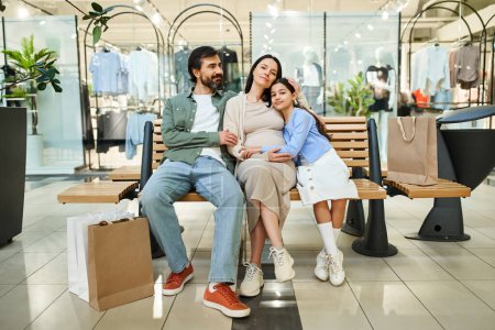 Téléchargez les photos : Une femme enceinte et sa fille partagent un moment tendre assis sur un banc dans un centre commercial animé. - en image libre de droit