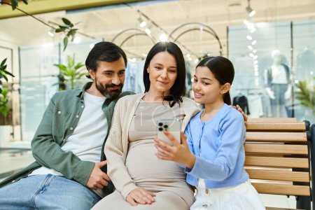 Foto de Una familia feliz de cuatro personas está sentada en un banco, absorta en la pantalla de un teléfono celular.. - Imagen libre de derechos