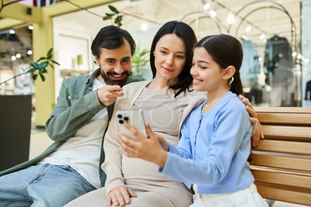 A happy family sits on a bench, engrossed in a cell phone together, enjoying a shopping weekend in the mall.