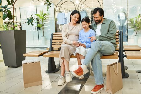Téléchargez les photos : Une famille heureuse assise ensemble sur un banc dans un centre commercial animé, profitant d'un moment de repos et de convivialité. - en image libre de droit