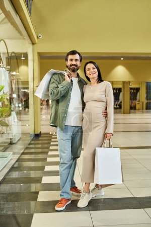 A pregnant couple happily holds shopping bags in a busy mall, enjoying a weekend outing together.