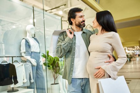 A pregnant couple joyfully holding shopping bags while strolling in a bustling shopping mall.