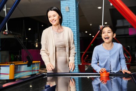 A mother and her daughter joyfully playing air hockey in an arcade during a fun weekend outing at the mall.