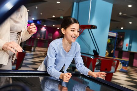 A mother and daughter enthusiastically play a game of air hockey, immersed in laughter and competitiveness at a mall gaming zone.