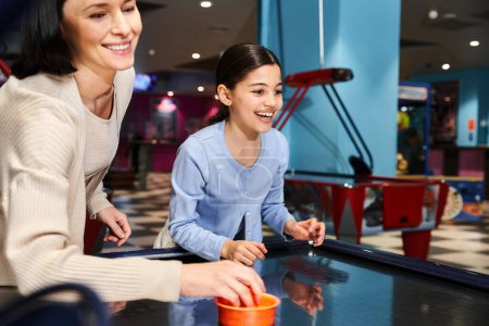 A joyful mother and daughter immerse themselves in a spirited air hockey match at a gaming zone in a mall on the weekend.