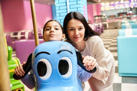 Téléchargez les photos : Une femme et un enfant joyeux posant ensemble à côté d'un jouet de carrousel coloré dans une zone de jeu de centre commercial. - en image libre de droit