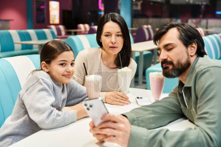 Foto de Una familia alegre se sienta junta en una mesa, absorta en mirar un teléfono inteligente, vinculación y pasar tiempo de calidad. - Imagen libre de derechos