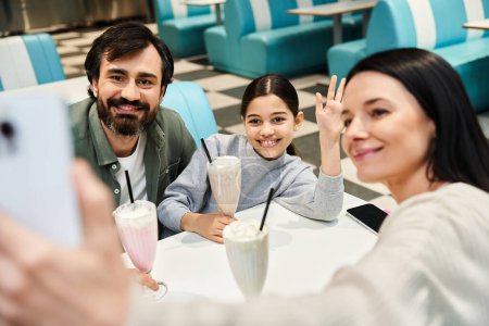 Téléchargez les photos : Une famille joyeuse capture un moment ensemble, souriant alors qu'ils prennent un selfie dans un charmant restaurant rétro pendant leur sortie de week-end. - en image libre de droit