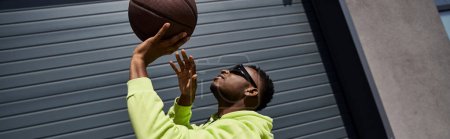 Téléchargez les photos : Un jeune homme afro-américain élégant attrape élégamment un ballon de basket en plein air. - en image libre de droit