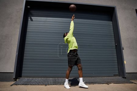 Stylish African American man dribbling basketball in front of garage.