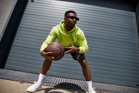Foto de Un hombre afroamericano con estilo en una sudadera con capucha verde sosteniendo una pelota de baloncesto. - Imagen libre de derechos