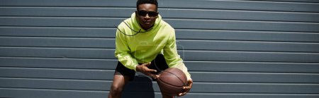 Schöner junger afroamerikanischer Mann in grünem Kapuzenpullover, der einen Basketball hält.