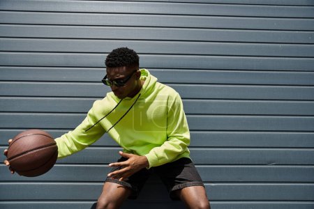 Téléchargez les photos : Jeune homme d'origine africaine dans un sweat à capuche vert vibrant tenant un ballon de basket. - en image libre de droit