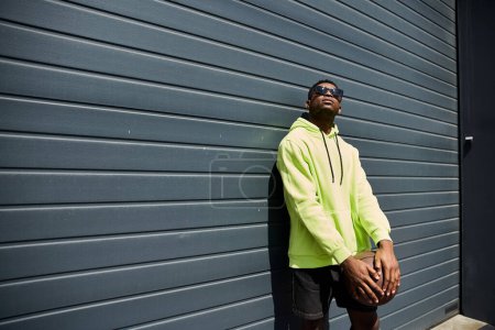 Stylish African American man leans against a wall in a yellow hoodie.