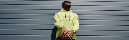 Handsome African American man leaning against urban wall in stylish yellow hoodie.