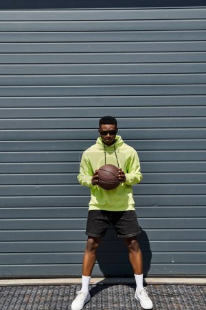 Fashionable African American man holding a basketball in front of a garage.
