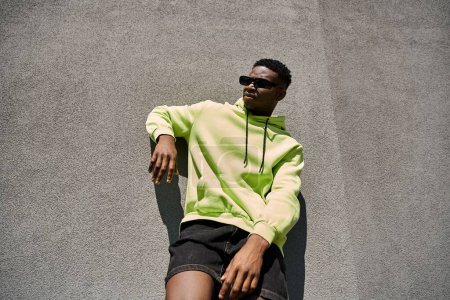 African American man in neon yellow hoodie and shorts, leaning against a wall.