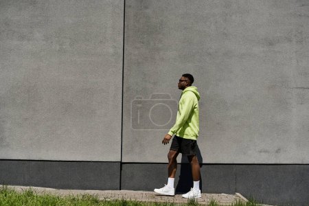Stilvoller afroamerikanischer Mann in neongrünem Kapuzenpullover läuft an einer Wand vorbei.