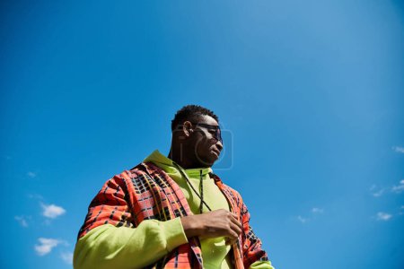 Stylish African American man in bright jacket stands under clear blue sky.