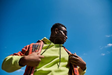 Téléchargez les photos : Un jeune homme afro-américain à la mode dans une veste jaune se tient devant un ciel bleu vibrant. - en image libre de droit