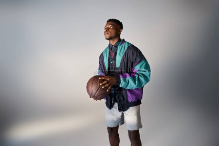 Téléchargez les photos : Beau Afro-Américain en tenue élégante tenant un ballon de basket sur un fond gris. - en image libre de droit