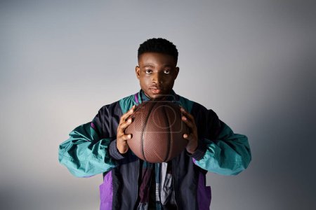bel homme afro-américain en tenue à la mode, tenant le basket-ball sur fond gris.