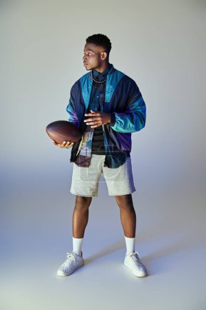 African American man in fashionable attire holding a football against white backdrop.
