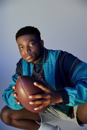 A stylish African American man crouching down while holding a football.