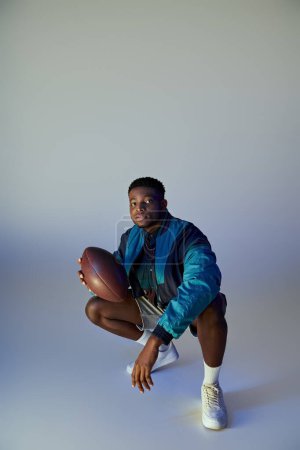 Téléchargez les photos : Bel homme afro-américain en tenue à la mode accroupi tout en tenant un football américain. - en image libre de droit