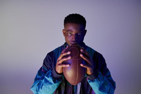 Elegante hombre afroamericano sosteniendo un balón de fútbol contra un fondo azul.