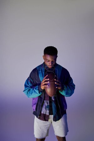 Photo for Young African American man in trendy outfit holding a football in front of blue background. - Royalty Free Image