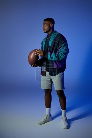 Téléchargez les photos : Un jeune Afro-Américain à la mode tenant un ballon de football devant un fond bleu. - en image libre de droit