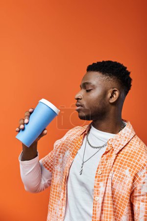 Foto de Hombre afroamericano guapo sorbe de la taza contra el vibrante fondo naranja. - Imagen libre de derechos